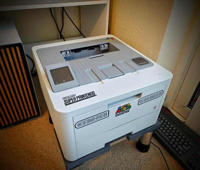 A beige and grey Brother laser printer modded with stickers to look like a Super Nintendo. The printer has a beige white color with a light gray rounded square area around the paper exit slot, much like the original Super Nintendo. A large "RESET" button sticker is placed on the bottom right corner of the gray area.
On the bottom left corner the Super Nintendo logo is applied as a sticker but it reads "Nintendo SUPER PRINTENDO".
On the front-facing side of the printer two large stickers on both sides are applied resembling the gamepad connectors. In-between, a colorful Super Mario logotype sticker is overlayed above the "brother" logotype, together it reads "Super Mario brother".