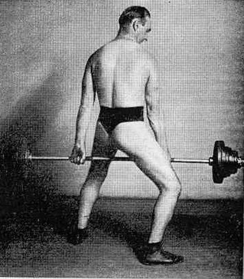 Black and white picture of Jefferson dressed only in thong, with the barbell lifted up between his legs in a quite awkward position.