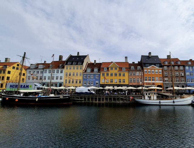 Nyhavn, the famous canal area in Copenhagen. The sky is pale blue, almost entirely covered by streaky white clouds. We are looking across the canal water to the iconic bank with its long row of historic, colourful terraced houses. Some three storeys, some four or five storeys, the gentle up-and-down variety of heights of the triangular rooftops gives the skyline a bit of a wing, like a line of musical notes. The houses are painted in a random variety of strong colours- light blue, dark blue, buttercup yellow, mustard yellow, deep orange, rust red. By contrast, the seawater in the canal is the colour of August in Denmark – cold hard black. On the opposite bank: people at pavement cafes, walking along - also some delivery trucks and cars because this is early morning. Two picturesque wooden boats are anchored facing one another, prow to prow, just below the wooden quay. One  is black. One is white, with Dannebrog, the Danish flag, waving off it. The flag is red with a white cross.