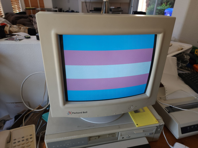A CRT on top of a 90s PC (with floppy disk) showing the Trans Pride Flag