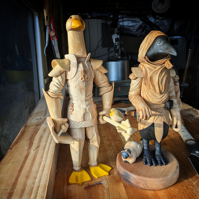 Two woodcarved humanoid birds. A goose knight in plate armor, with a shield and a flanged mace with a red robin sitting on it. The other sculpture is a crow with its right hand on the handle of its sword. A squirrel is climbing its left arm, a cat sits next to its feet.