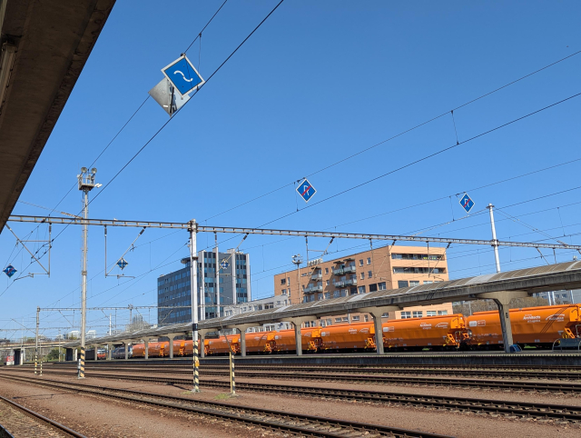 A view of a railway station with overhead wires. In the wires hangs a sign with white sinusoid on a blue background as well as a few similar signs with crossed letter R.
