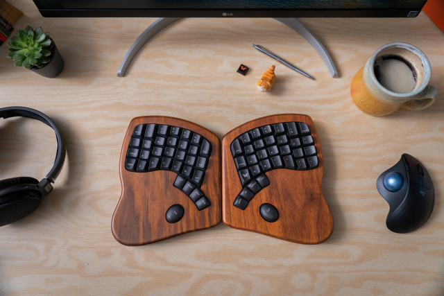 A "split" keyboard sitting on a desk. The base of the keyboard is an attractive dark-brown wood, and the keys are black, with deep curved divots on top. The keyboard is split into two pieces -- the left-hand part of the QWERTY key layout on the left, and the right-hand part on the right. There is also a trackball mouse, cup of coffee, and set of headphones next to it. Overall it looks like a pretty sweet, de luxe and ergonomic workstation!