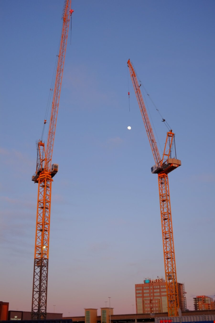 Two cranes in front of the moon appearing in the still blue sky.
