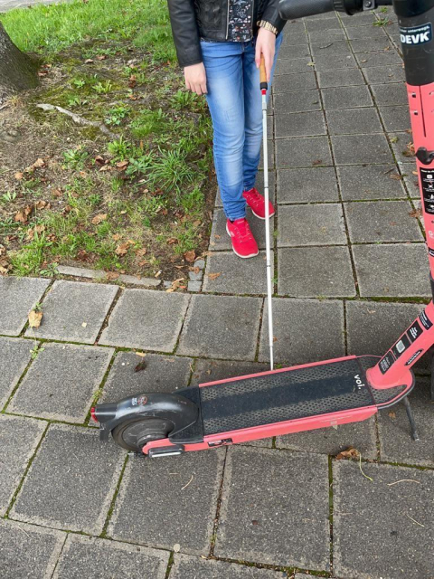 Eine Person, die ihren  Blindenlangstock benutzt, steht auf einem Gehweg vor einem quer abgestellten E-Roller. Dieser blockiert fast die gesamte Gehwegbreite. Die Person ist ungefähr bis zur Brust zu erkennen,  trägt eine schwarze, offene  Lederjacke, mittelblaue Jeans mit helleren Stellen und auffällig rote Turnschuhe. In der linken Hand hält sie den  weißen Langstock mit roter, runder Spitze, der  unter dem Trittbrett des E-Rollers verschwindet.
Der Roller ist rot mit schwarzen Elementen, gehört zum Anbieter VOI und trägt am Lenker einen DEVK-Sticker. Der Gehweg besteht aus grauen rechteckigen Betonplatten. Links daneben verläuft eine schmale Rasenfläche mit Moos, Grasbüscheln, Erde und trockenen Blättern. Die Szene ist bei Tageslicht aufgenommen. Das Bild ist scharf, die Farben sind kräftig. Der abgestellte Scooter stellt eine klare Barriere auf dem Weg dar und erschwert die ungestörte Fortbewegung für die blinde Person.