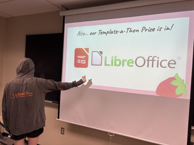 An individual stands house left to a projector at the front of a university classroom, facing away from the camera. The individual is seen wearing a grey hoodie with the LibreOffice logo in orange on the back, and is holding up their right arm, making a heart symbol with their thumb and forefinger. On the projector is a slide of a slide show with the LibreOffice and LibreOffice Impress logos and text that reads, “Also… our Template-a-Thon Prize is in!”