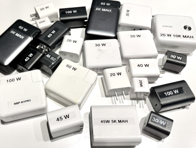 collection of usb chargers on a table, some black, some white, each with a prominent diy label stuck on listing the wattage of the charger.