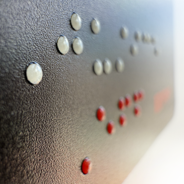 Tactile graphic sticker depicts figure of Lady Justice with fiery hair and flowing garment below armor, holding a sword and scales, with braille reading ‘Justice’ in white, and ‘EFF’ in red. The figure’s body, legs, arms, hair, and objects are raised, showing the outlines tactually so you can show off and feel the design.