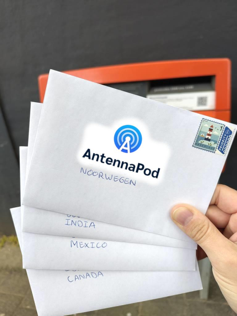 Photo of four small envelopes held in hand, in front of an orange post box, with a dark grey wall behind it. The envelopes have the countries written visibly in Dutch: Norway, India, Mexico and Canada. The top envelope has a 'priority' stamp and the recipient name and address obfuscated by the AntennaPod logo.