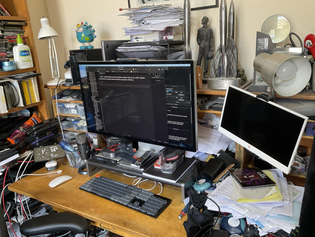 An author's writing bureau, bearing monitors, keyboard, huge amounts of paper, and a few Hugo awards tucked away at the back.