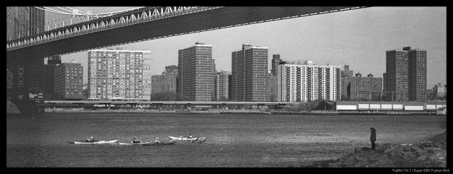 A person stands on a rocky part of the bank of the East River.  They are looking at a group of kayaks paddling past, both beneath the Manhattan Bridge.  The New York City skyline is visible in the background.