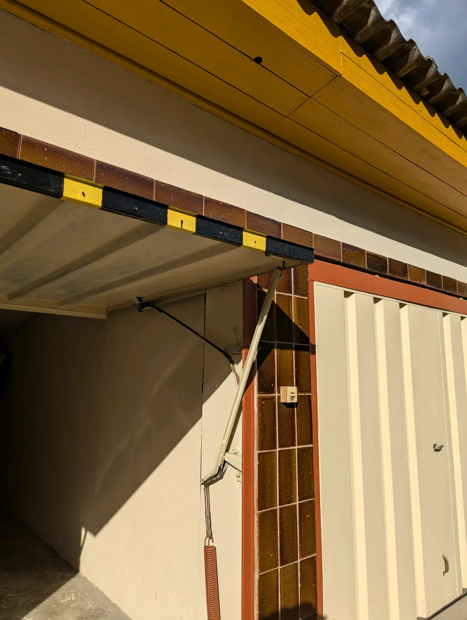 Zwei Geragentore. Eines ist hochgeschoben und man sieht an der Stirrnseite eine farbige gelb-schwarze Signalschraffierung. Das andere ist geschlossen und in hellbeschem Ton gestrichen. Der Dachkasten ist gelb/oker. Steinmehl und Leinöl sind die Komponenten der Farbe.