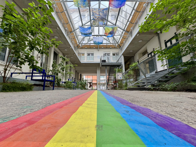 A picture taken from the ground, within a venue hall. On the ground there is a long Pride flag. On the ground floor, the hall is surrounded by greenery. Upstairs, the hall is surrounded by balconies. 