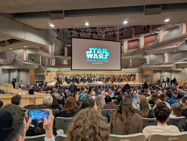 A scene from the back of a crowded auditorium.  In the front and center, quite high up, is a large front projection screen displaying the title screen "Star Wars" / "The Empire Strikes Back" / "In Concert".

Below the screen is a full orchestra, ready to fill the room with John Williams's best.