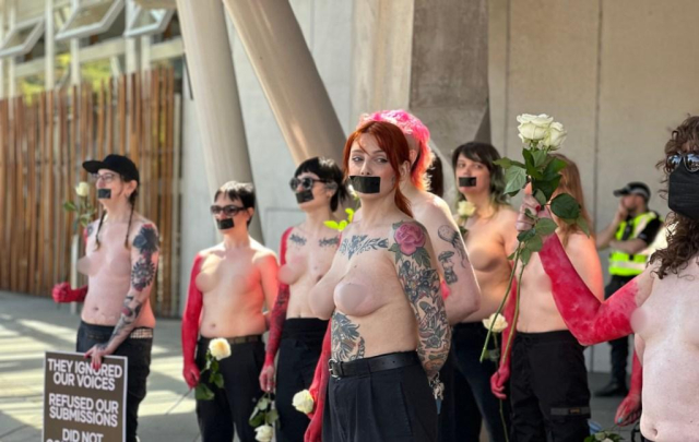 8 topless women with black masking tape over their mouths, in front of a building. A police officer is monitoring behind them. The photograph has been censored to obscure breasts and/ or nipples.