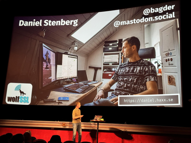 Daniel on stage in front a huge display showing a slide with Daniel himself on