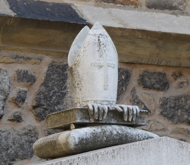 Detail of an episcopal monument in Brno: a mitre sitting on a book, with two gloves protruding from beneath the mitre and nervously gripping the side of the book.