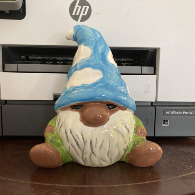 a colourful ceramic garden gnome with a blue hat featuring cloud designs, a white beard, and green pyjamas, sitting on a wooden surface near an hp printer