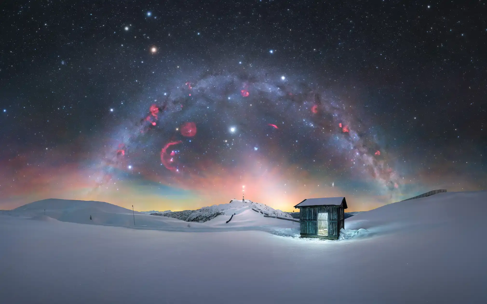 A panorama of the winter Milky Way with reddish nebulae, stretching above Dobratsch.
