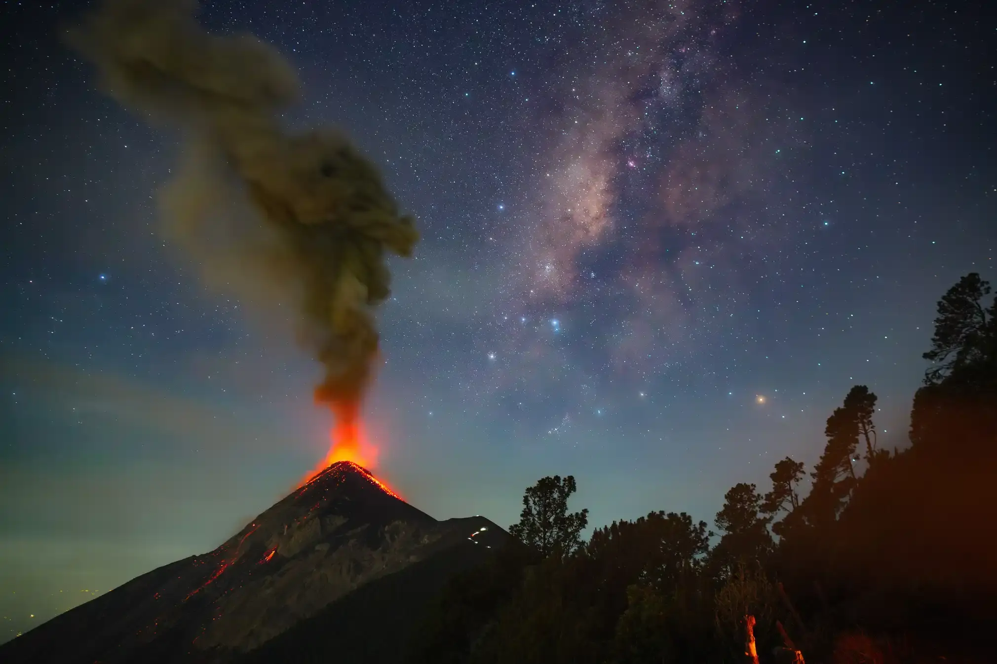An active volcano. Above, the Milky Way stretches diagonally across the sky. As the volcano erupted, the ash plume rose vertically, forming an acute angle of about 45 degrees with the galaxy’s diagonal path, creating a stunning visual contrast between Earth’s fury and the cosmos’ serenity.