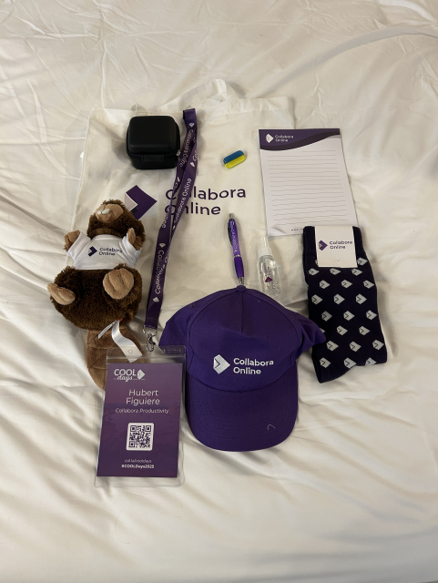 A few items spread on a white bedding. Most of them are branded Collabora Online. Included a white tote bag, an Ukraine pin, a black device, a beaver plush, a purple baseball cap a purple pen, socks, a bottle of hand sanitizer and a notepad. Also a conference badge.