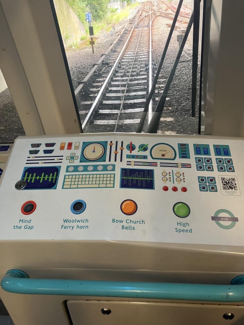 The front seat of the DLR; the top of the locked access has a sticker on top for kids to play at being the train conductor, with colourful painted dials, buttons, and switches. Four buttons are labelled: Mind the Gap, Woolwich Ferry horn, Bow Church Bells, High Speed.