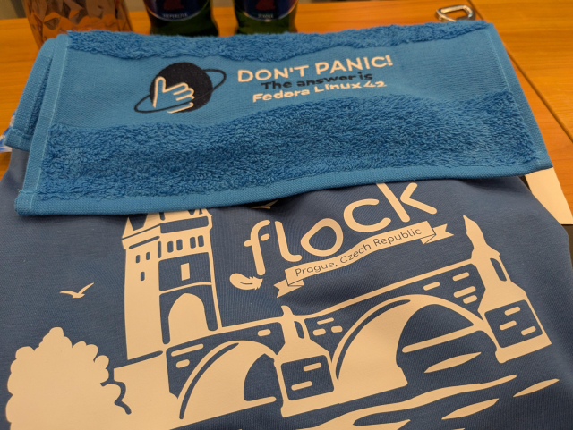 A blue t-shirt with the Charles Bridge and Flock and a blue towel which says: Don't panic, the answer is Fedora Linux 42.