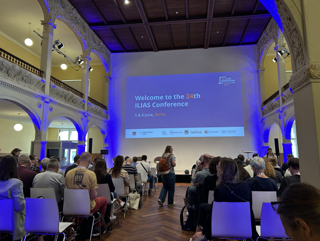 Picture of an auditorium with a projection screen showing a slide saying “Welcome to the 24th ILIAS conference”. To the top left there is a mezzanine. An attendee is looking in the aisle looking for a seat.