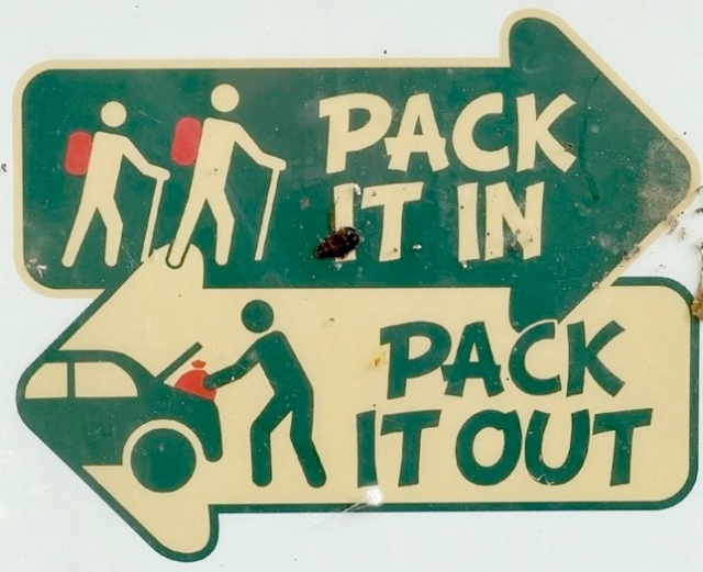 Sign with two arrows: one pointing right showing hikers with backpacks and the text "Pack it in," and one pointing left showing a person loading a car with the text "Pack it out."
