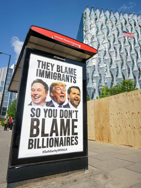 olitical street poster outside the U.S. Embassy in London featuring edited images of Elon Musk, Donald Trump, and another billionaire, with bold text reading: “They blame immigrants so you don’t blame billionaires.” The poster includes the hashtag #SolidarityWithLA, referencing ongoing protests in Los Angeles. The striking image juxtaposes the sleek U.S. Embassy architecture and American flag with a grassroots message challenging wealth inequality and scapegoating.