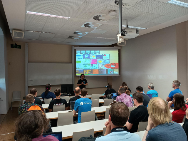 Room with people on chairs. One person in the front, behind him a slide with CD and DVD covers of GNU/Linux distributions.