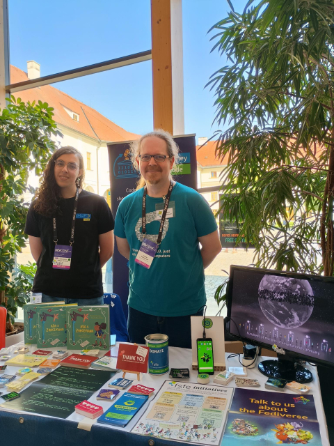 Two people behind a desk full of flyers, stickers, and books and a screen showing a video. In the background some plants.