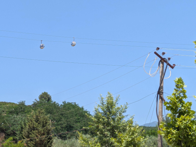 A view of a blue sky. There are two gondolas of a cable car high in the sky. There is also a wooden pole supporting three wires apparently of a high voltage line.