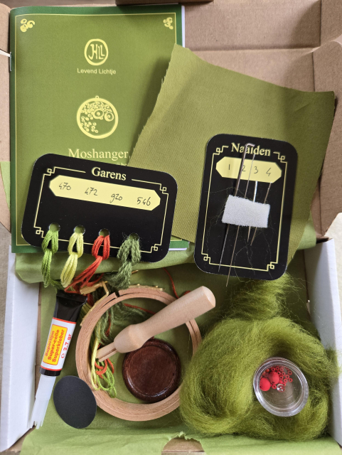 Small box, opened to show the contents: moss green wool roving, embroidery yarns, embroidery needles, a needle felting needle with holder, an embroidery ring, a piece of moss green fabric, a small wooden disk with a decorated rim, super glue, small red beads and hand made clay red mushrooms
