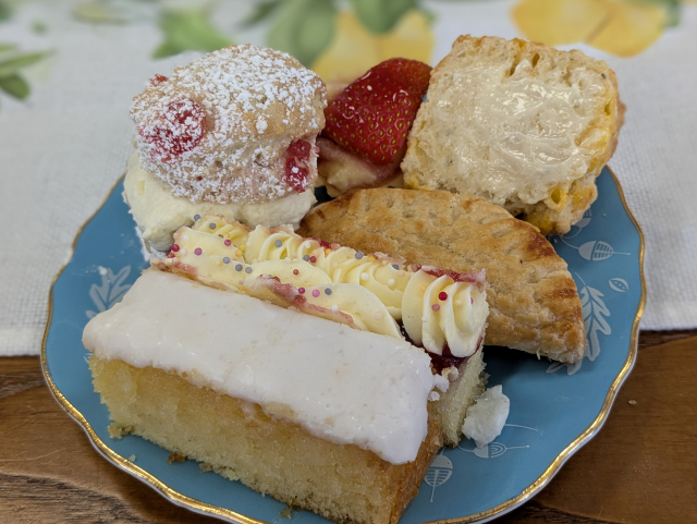 Colour photograph of a pale blue vintage tea plate edged with gilt. It is crammed with small cakes and scones.