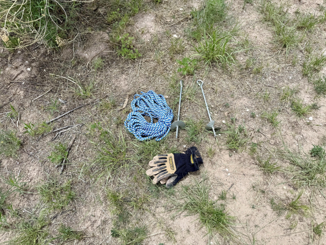 The image shows a collection of items laid out on a patch of ground with sparse grass and dirt. In the center, there is a coiled blue and white rope, suggesting it is used for securing or lifting. To the right of the rope, there are two metal stakes with eyelets at the top, likely used for anchoring the rope into the ground. Below the rope, a pair of black and tan gloves is visible, indicating they are used for manual work or protection. The ground is uneven with patches of grass and dirt, and there are some small twigs scattered around. The overall setting appears to be outdoors, possibly in a garden or construction area.

Provided by @altbot@fuzzies.wtf, generated privately and locally using Ovis2-8B

🌱 Energy used: 2.574 Wh
