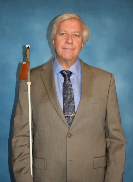 Image of Jeff Altman standing with his white cane by his side, in front of a blue background. He is wearing a suit and tie.