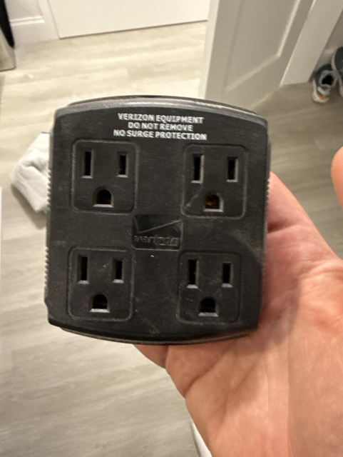 A black four outlet block that reads "VERIZON EQUIPMENT DO NOT REMOVE NO SURGE PROTECTION", removed, in my hand. 