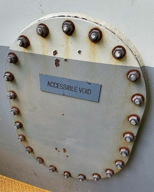 A metal hatch closed by a zillion rusty bolts has a label on it that reads "accessible void" 