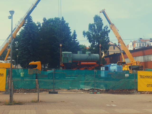 Na stavenisti stoji vedle sebe dva vysoke zlute jeraby, ktere zvedaji starou parni lokomotivu se zelenym telem a cervenym podvozkem do vzduchu.
