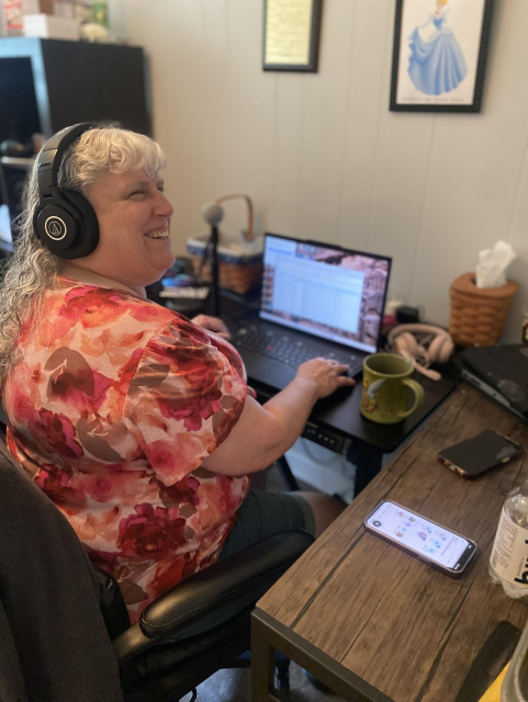 Photo of Cindy Hollis seated in front of her laptop at her home office. Cindy wears headphones and her phone sits next to her on the desk, along with a cup of coffee. Cindy is smiling towards the camera. 