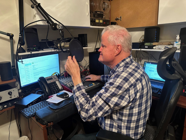 Photo of Larry Gassman seated in his home office, which is a professional recording studio. Larry is seated at a desk with multiple computer monitors, audio equipment, and broadcasting gear. He's positioned in front of a professional microphone.  