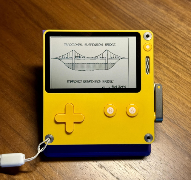 Playdate on a desk, open to a "suspension bridge" xkcd comic