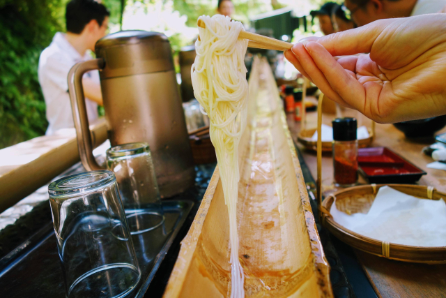 Person holding a clump of cold noodles over a half bamboo slide in a restaurant setting 