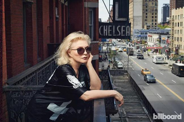 Deborah Harry on a balcony wearing sunglasses in front of the Hotel Chelsea sign.