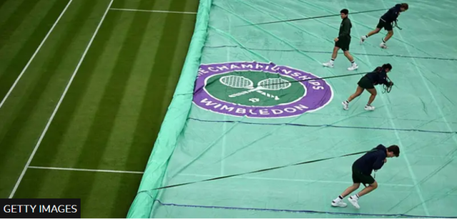 a group of people on a grass tennis court. They are covering the court with a large green tarp. The tarp features a circular purple and white emblem in the center, displaying two crossed tennis rackets and a tennis ball, with the words "The Championships Wimbledon" around it. Four individuals are seen walking on the tarp, each pulling the cover into place. The grass is neatly striped, and there are visible lines marking the boundaries of the tennis court.