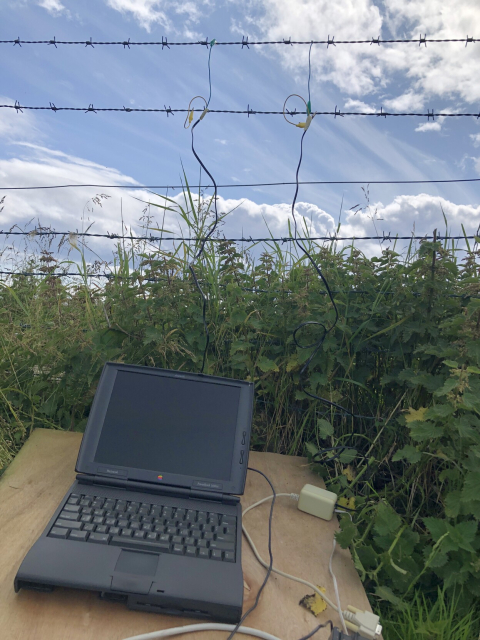 An old Mac laptop with a barbed wire fence behind it, and some suspicious cables clipped to the barbed wire