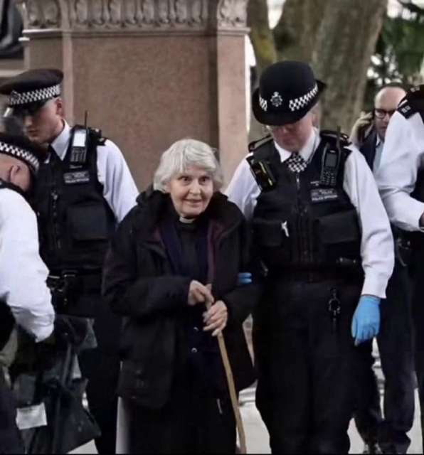 The picture shows a white-haired woman wearing a dark jacket over a priest's vestments (with color visible) and carrying a wooden cane. She is being escorted by a police officer, while other police officers are visible in the background.
