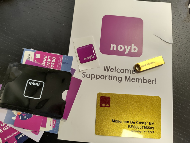 A collection of noyb supporting member items is arranged on a dark tabletop. A large white card shows a purple square logo and the text 'Welcome Supporting Member!'. A glittery gold membership card reads 'Molleman De Coster BV BE0860796509 Member N° 7224'. A small gold USB drive is engraved 'GOLD MEMBER'. A square purple sticker says 'noyb' and a black cardholder bears a white noyb logo among assorted purple and pink promotional flyers. 
