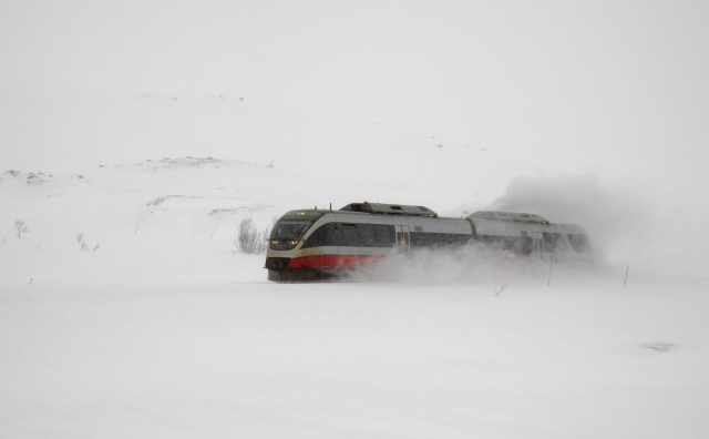 Bombardier Talent train in older NSB livery, covered in a cloud of snow as it zooms through snow-covered Nordlandsbanen